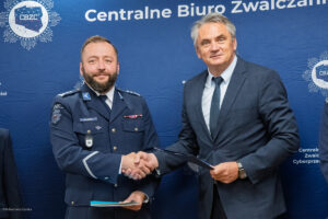 Dwóch mężczyzn stoi na tle niebieskiego baneru z napisem "Centralne Biuro Zwalczania Cyberprzestępczości". Jeden z nich, po lewej stronie, jest ubrany w mundur policyjny z odznakami i naszywkami, a drugi, po prawej stronie, ma na sobie elegancki garnitur. Obaj mężczyźni podają sobie ręce i patrzą w stronę aparatu. Baner w tle zawiera również logo z literami "CBZC".